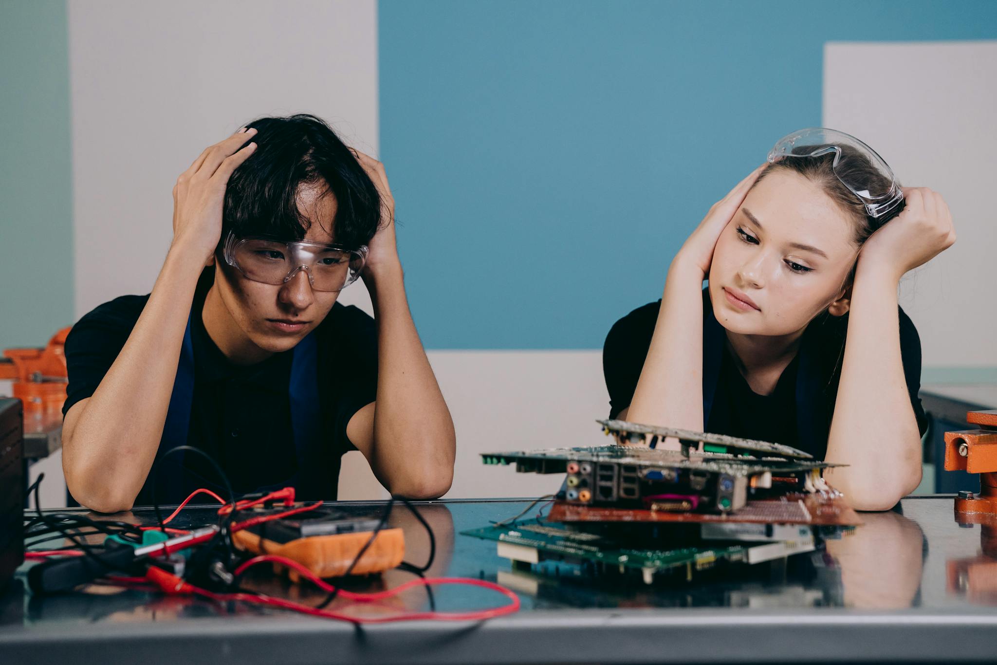 Two technicians in safety goggles work on circuit boards, displaying stress and focus in a tech lab.